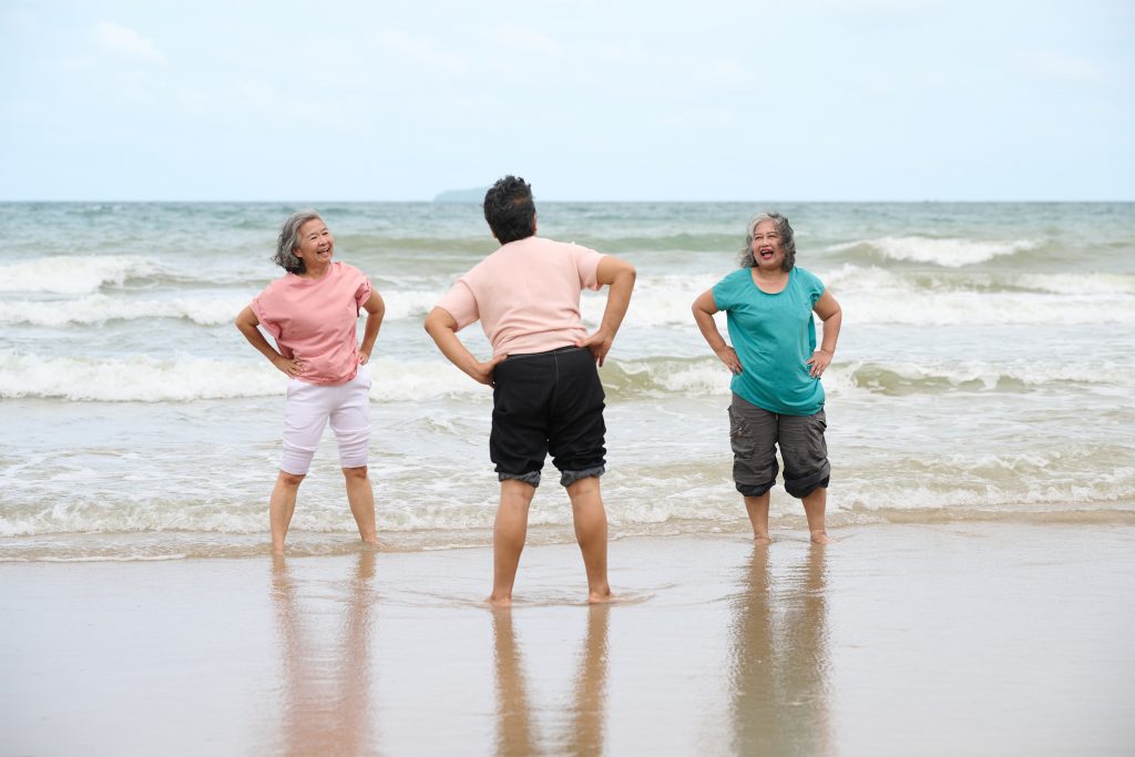 Elderly women exercising on the beach, with gentle waves lapping at their feet. The two women on the sides are stretching