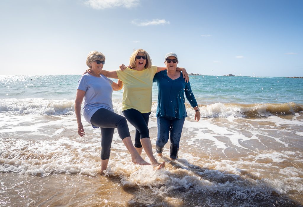 Lovely group of senior girl friends on their 60s walking and having fun splashing water on sea. Three mature healthy retired females Laughing and enjoying retirement and outdoors active lifestyle.